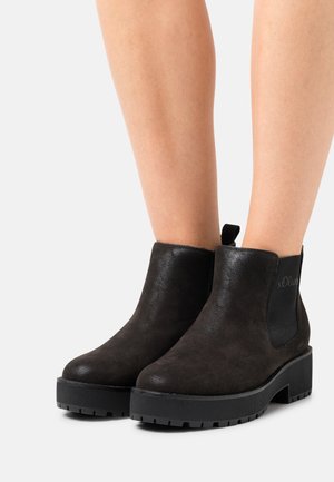 Schwarze Wildleder-Ankle-Boots mit einem klobigen Absatz, elastischen Seiteneinsätzen und einem Logo-Detail an der Außenseite. Strukturiertes Profil für besseren Halt.