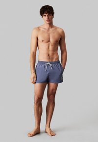 Pantaloni da bagno da uomo in materiale blu scuro, con un laccio bianco e un logo ricamato sul lato sinistro. Elastico in vita.