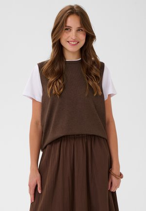 Femme souriante aux longs cheveux bruns porte un gilet en laine marron sur un t-shirt blanc et une jupe marron, debout devant un fond uni.