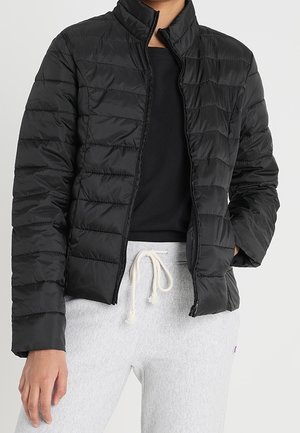 Person iført en sort quiltet jakke med lynlås, sort top og lysegrå joggingbukser med hvid snor.