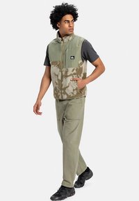 Jauns vīrietis, tērpies zaļā kamuflāžas vilnas vesti, tumši pelēkā T-kreklā, gaiši zaļās biksēs un melnās sporta kurpēs, staigā ar vienu roku kabatā.