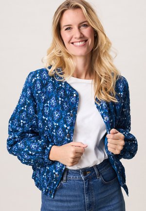 Lachende vrouw met lang blond haar, gekleed in een blauw gewatteerd jack met patroon, een wit overhemd en een blauwe spijkerbroek, houdt de voorkant van het jack vast.