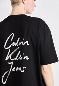 Svart t-skjorte laget av bomull med stor hvit grafisk tekst på ryggen som sier "Calvin Klein Jeans." Rund hals, avslappet passform.