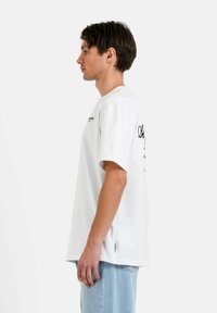 Camiseta blanca de manga corta con cuello redondo. Presenta texto impreso en el pecho y la espalda. Se lleva con unos pantalones vaqueros de denim azul claro.