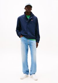Marineblaues Leichtgewicht-Jackett mit hohem Kragen, grünem T-Shirt darunter, hellblauen Jeans und weißen Sneakers. Auch eine hellblaue Mütze gehört dazu.