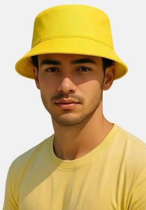 Jonge man met getrimde baard die een felgele bucket hat draagt en een bijpassend geel T-shirt tegen een effen achtergrond.