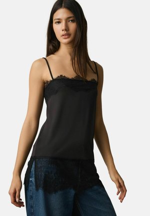 Woman wearing a black sleeveless lace-trimmed top and blue jeans, standing against a plain white background.