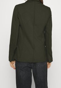 Mujer vistiendo un blazer a medida de color verde oliva y jeans de mezclilla negros, de pie con los brazos relajados a los lados, de espaldas a la cámara.