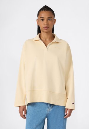 Donna con i capelli intrecciati che indossa una polo beige lunga a maniche lunghe e jeans blu, in piedi davanti a uno sfondo chiaro e uniforme.