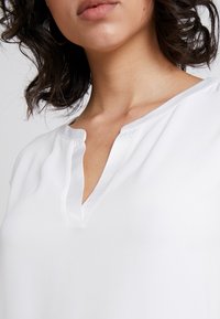 White blouse with a V-neckline and short sleeves, made of soft fabric. Features a subtle sheen and relaxed fit for comfort.