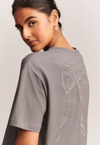 Camiseta gris de tela suave, con un diseño de lazo plateado brillante bordado en la espalda, con cuello redondo y mangas cortas.