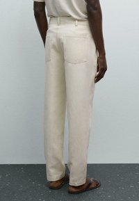 Beige, high-waisted trousers with a loose fit, featuring two back pockets and stitched detailing. Made from cotton material.