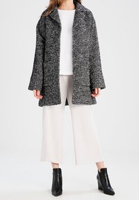 Cappotto in lana bianco e nero a trama con grande colletto, due bottoni frontali e tasche a soffietto, abbinato a una maglia bianca e pantaloni a gamba larga.