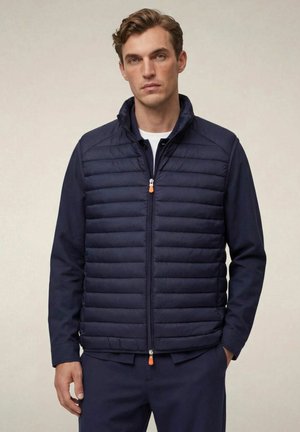 Giovane uomo che indossa una giacca trapuntata blu navy con cerniera sopra una camicia a maniche lunghe, in piedi con una mano in tasca su uno sfondo uniforme.