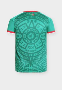 Zelený športový dres s červenými a bielymi lemami na rukávoch, s veľkým dizajnom aztéckeho kalendára a textom "Somos México" blízko goliera.