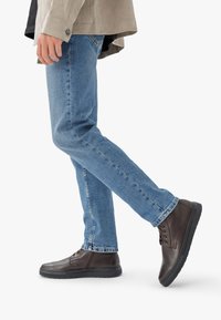 Botas de cuero marrón hasta el tobillo con puntera redonda, suela de goma negra y diseño de cordones, combinadas con jeans azules y una chaqueta de color claro.