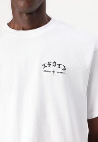 Balta kokvilnas T-kreklu ar melnu grafisko druku, kurā ir Japāņu rakstzīmes un teksts "EDWIN SUPPLY" kreisajā krūšu daļā.