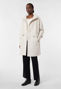 Cappotto esterno in misto lana color avorio con bottoni a pressione, grandi tasche e dettagli a coste sulle maniche. Abbinato a pantaloni neri e stivaletti.