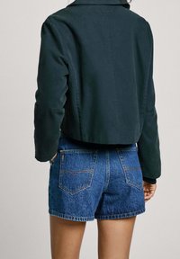 Ciemnozielona krótka kurtka z gładkiego materiału, z kołnierzykiem koszulowym. Połączona z jasnoniebieskimi szortami dżinsowymi, z widocznymi tylnymi kieszeniami.