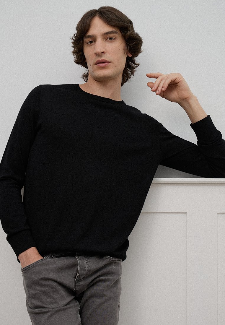 Schwarzer Pullover mit langen Ärmeln und rundem Halsausschnitt, weicher Textur und gerippten Bündchen, kombiniert mit grauen Slim-Fit-Jeans, die an eine Wand gelehnt ist.