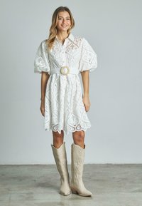 Biała koronkowa sukienka z bufkami, z wyszczuploną talią dzięki pasowi z okrągłą klamrą, zestawiona z beżowymi kowbojkami sięgającymi do kolan.
