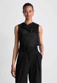 Melns bez piedurknēm tops ar drapētu kakla izgriezumu, apvienots ar augsti jostas melniem biksēm, kurām ir priekša pleats un sānu kabatas.