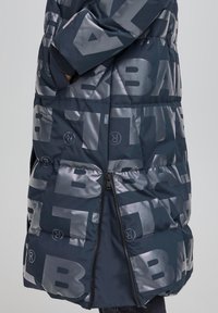 Chaqueta acolchada azul marino con gran logotipo plateado, que cuenta con una abertura lateral con cremallera y puños acanalados. Tela lisa y brillante con textura acolchada.