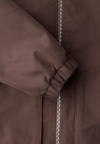Manche de veste marron avec un poignet élastique reposant sur l'avant de la veste à côté d'un détail de fermeture éclair argentée visible.