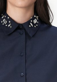 Camisa azul-escura com botões, apresentando um colarinho decorado com várias embellishments transparentes e metálicas. Textura de tecido suave.