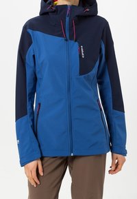 Personne portant une veste d'extérieur à capuche bleu et marine avec des poches zippées à l'avant et sur la poitrine, sur un pantalon marron.