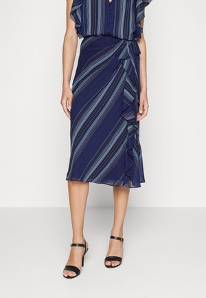 Mujer vistiendo una falda cruzada azul marino con rayas diagonales en azul claro y detalle de volantes, combinada con sandalias negras de tacón alto.