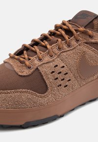 Scarpa atletica marrone con tomaia in suede texturizzato, pannelli in mesh, suola in gomma e lacci marroni a contrasto. Presenta perforazioni distintive.