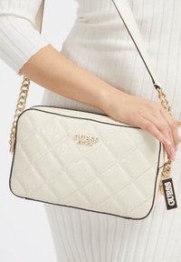 Beige quiltet håndtaske med guldbelægning, præget logo og justerbar kædestrop. Har sorte detaljer og et mærke med brandnavnet.