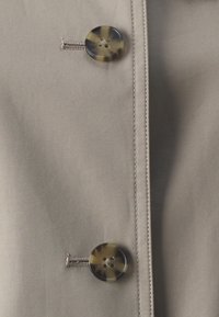 Jasno beżowa tkanina z przekątnym ryblem; zawiera dwa okrągłe guziki w kolorze tortoise z czterema otworami, przyszyte srebrną nitką.