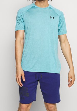 Mann mit hellblauem kurzärmligem Sportshirt und dunkelblauen Under Armour Shorts, steht vor einfarbigem Hintergrund.