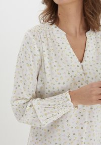 Vit blus med ett blommönster som har gula och rosa blommor. Mjuk, texturerad tyg med V-ringning och samlade ärmar.