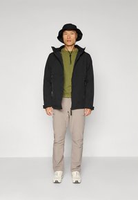 Zwart lichtgewicht jack met capuchon, groen shirt, beige broek en witte sneakers. Accessoires omvatten een zwarte bucket hat en een trekkoord.