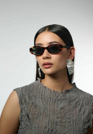 Des lunettes de soleil en écaille de tortue avec un design allongé et des verres sombres, associées à de grandes boucles d'oreilles en argent texturé. Le tissu gris plissé du haut est visible.