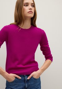 Fuchsia Pullover mit rundem Hals, gerippten Bündchen und Saum. Weicher Stoff, taillierte Silhouette. Kombiniert mit blauen Jeans.