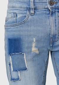 Jean en denim bleu clair avec des patchs usés, présentant trois textures de tissu différentes et plusieurs nuances de bleu, y compris des bords effilochés.