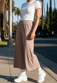 Personne portant un t-shirt blanc, un pantalon beige à jambes larges, des chaussettes blanches et des baskets blanches, debout sur un trottoir ensoleillé avec des palmiers à proximité.