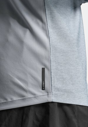 Primo piano di una maglietta atletica grigio chiaro con motivo a griglia sul lato sinistro, tessuto uniforme sul lato destro e etichetta nera "X-VENT" vicino all'orlo.