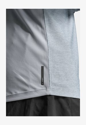 Primo piano di una maglietta atletica grigio chiaro con motivo a griglia sul lato sinistro, tessuto uniforme sul lato destro e etichetta nera "X-VENT" vicino all'orlo.