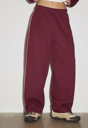 Personne portant un pantalon de survêtement bordeaux ample avec des bandes blanches sur les côtés et des chaussures de randonnée beiges, debout sur un sol gris contre un mur blanc.