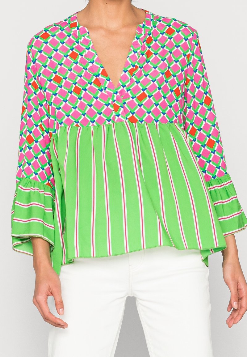 Blouse colorée avec un motif géométrique rose et vert, col en V et manches évasées. La moitié inférieure présente des rayures vertes avec des accents blancs.