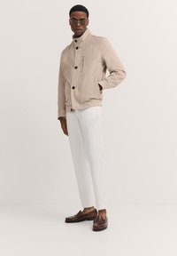 Beige suede jacket with a stand collar, button closure, and a zip pocket. Paired with white trousers and brown loafers, featuring smooth textures.