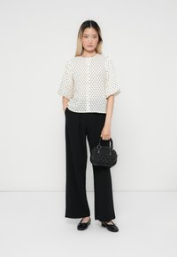 Blouse blanche à pois avec des manches bouffantes, associée à un pantalon noir à pattes d'éléphant et un petit sac à main noir texturé. Des chaussures plates noires complètent la tenue.