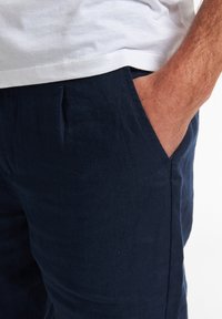 Pantaloni in lino blu navy con una vestibilità rilassata, dotati di tasche laterali e una superficie testurizzata. Il tessuto appare leggero e traspirante.