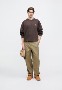 Sweatshirt maron cu un logo mic, pantaloni verzi măslinii, pălărie bej în mână și cizme portocalii, stând pe un fundal alb simplu.