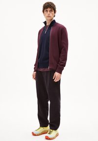 Un cardigan zippé bordeaux sur un pull marine, associé à un pantalon en velours côtelé noir et des baskets jaunes vives avec des accents orange.